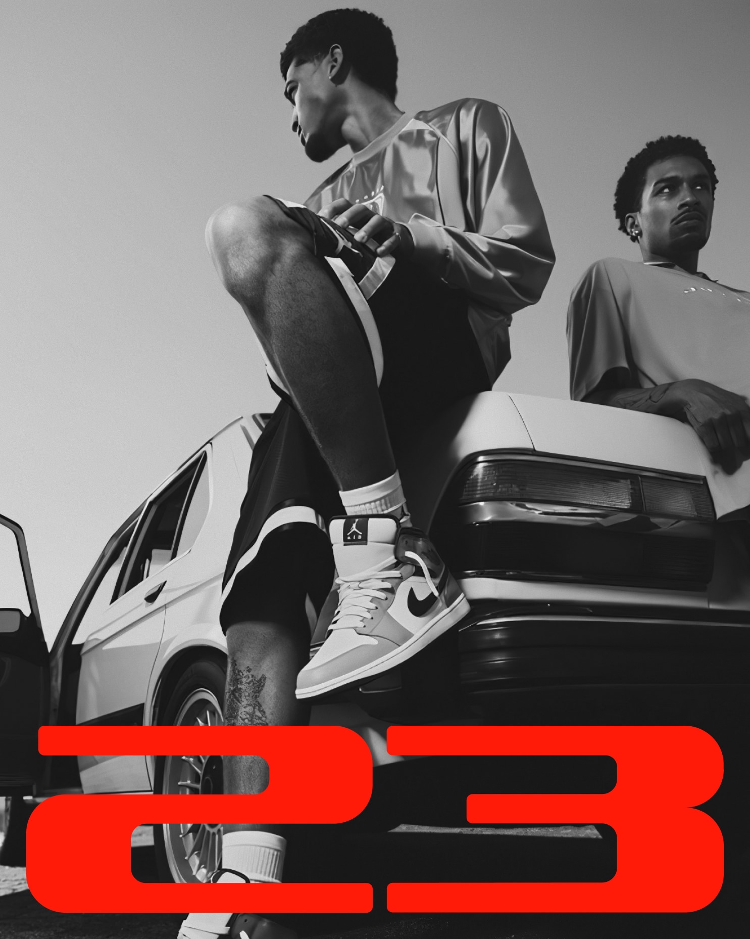 Black and white photo of two men beside a vintage car, one wearing Jordan 1 sneakers, with large red "23" wordmark overlaid at bottom